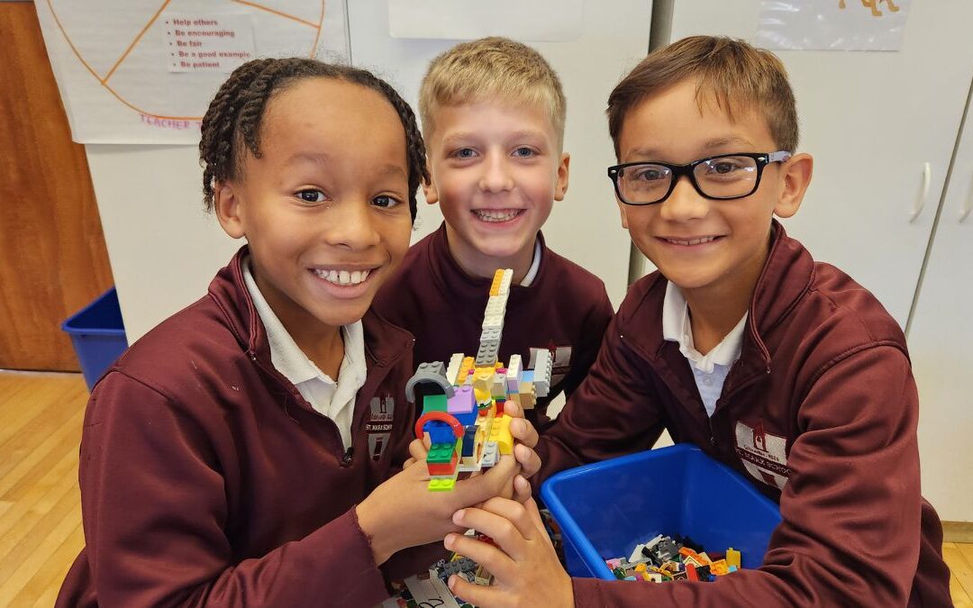 Three St. Mark School students smile and hold up a LEGO creation together in a classroom.